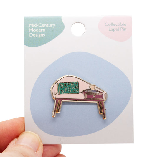 Mid-century modern bench enamel pin. gold plated with pink, teal and deep pink enamel. Pin is on matchbook backing card, held by hand, over white background.