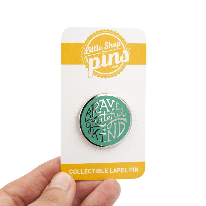 silver enamel pin with greenish teal enamel fill. Says, "Brave, grateful, kind". Pin is on a backing card, being held by a hand, over a white background.