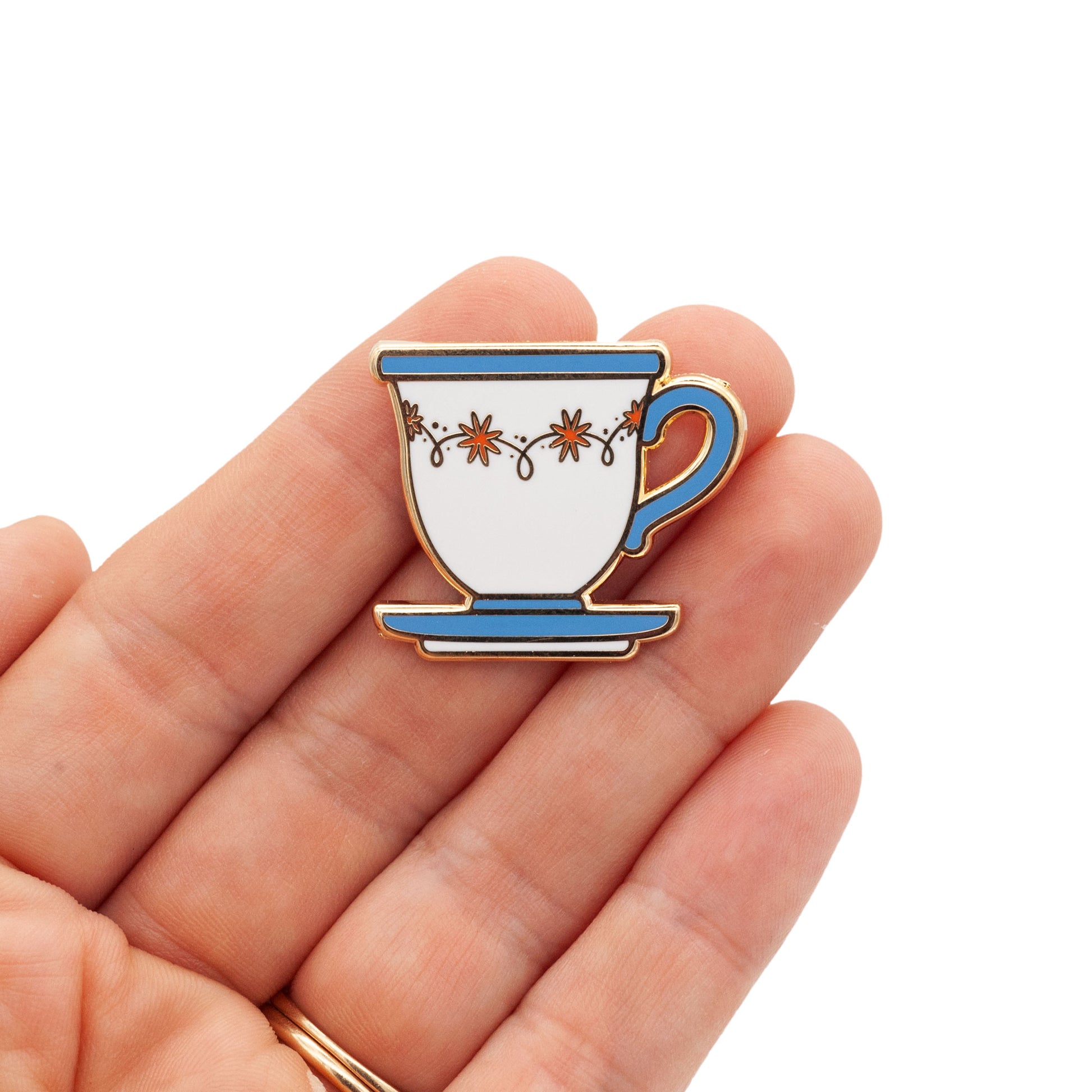 An illustrated teacup in pin form. The teacup has a handle on its right side that is blue. Colors being used are blue, red and white. gold plated and hard enamel. the pin is being held by a hand against a white background.