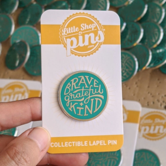 silver enamel pin with greenish teal enamel fill. Says, "Brave, grateful, kind". Pin is on a backing card, being held over a bunch of other pins.