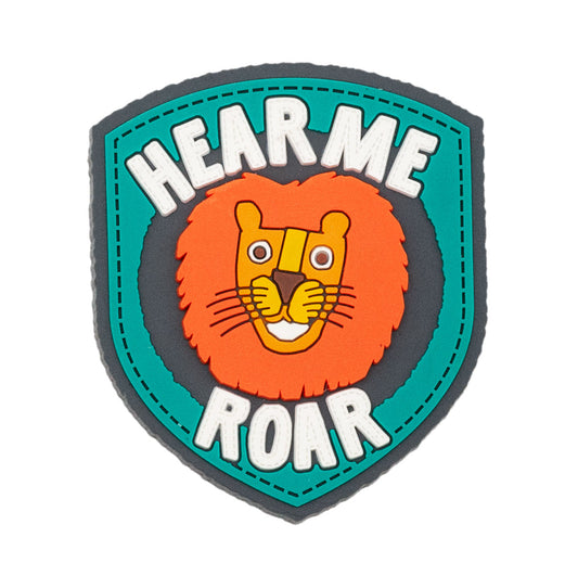 Lion face, smiling surrounded by type saying "Hear me Roar." Soft PVC made of colors: teal, dark teal, orange, brown, yellow, and white.