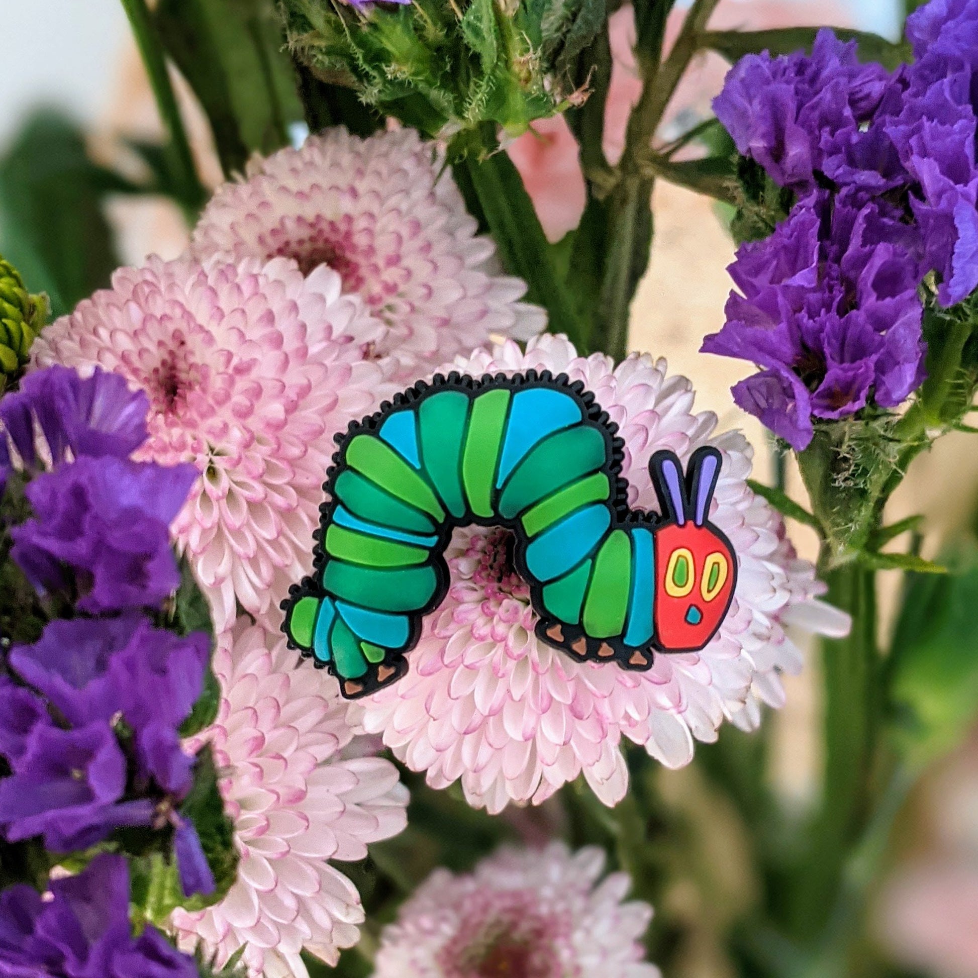 The Very Hungry Caterpillar PVC enamel pin on white background. Caterpillar has a red face with yellow and green eyes , with purple antennas, and a light green and dark green body. the pin is sitting on a pink flower and is surroundsed by other purple and pink flowers.