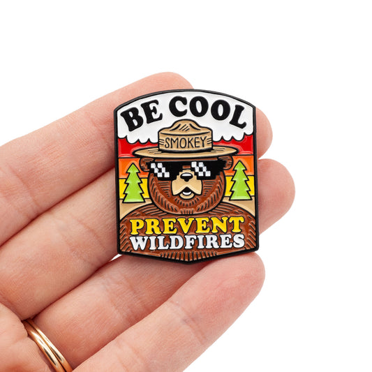 smokey bear with cool sunglasses on and a forest background. type says "be cool, prevent wildfires". Pin is being held by hand, over white background.