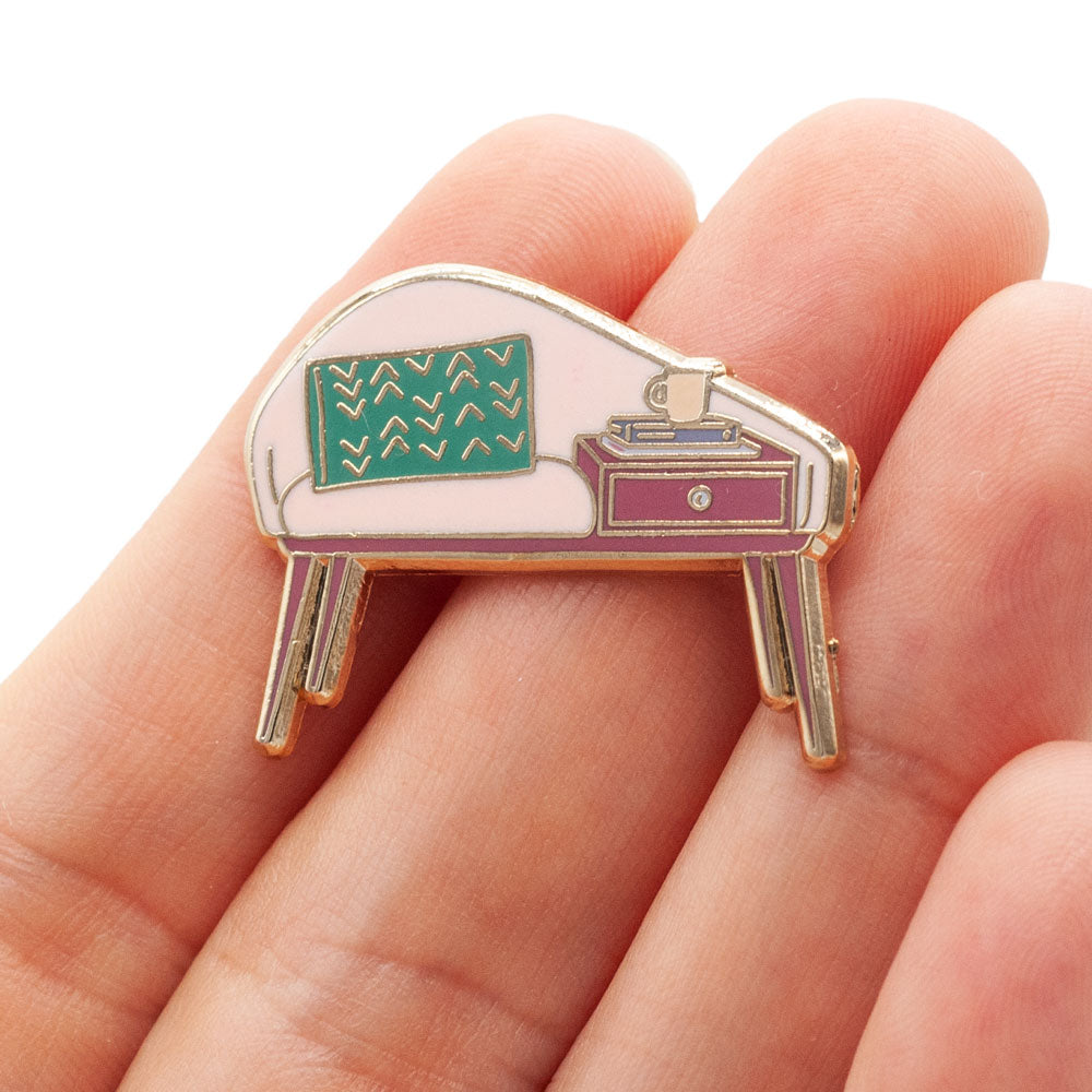 Mid-century modern bench enamel pin. gold plated with pink, teal and deep pink enamel. Pin is being held by a hand, over white background.