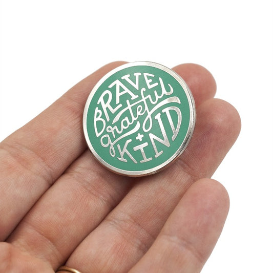 silver enamel pin with greenish teal enamel fill. Says, "Brave, grateful, kind". Pin is held by a hand over white background.
