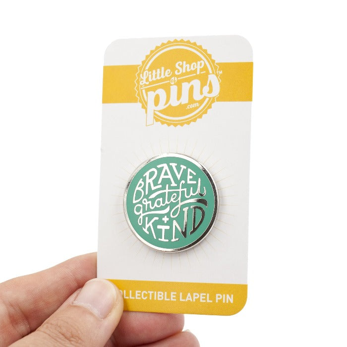 silver enamel pin with greenish teal enamel fill. Says, "Brave, grateful, kind". Pin is on backing card, being held by hand, over white background.