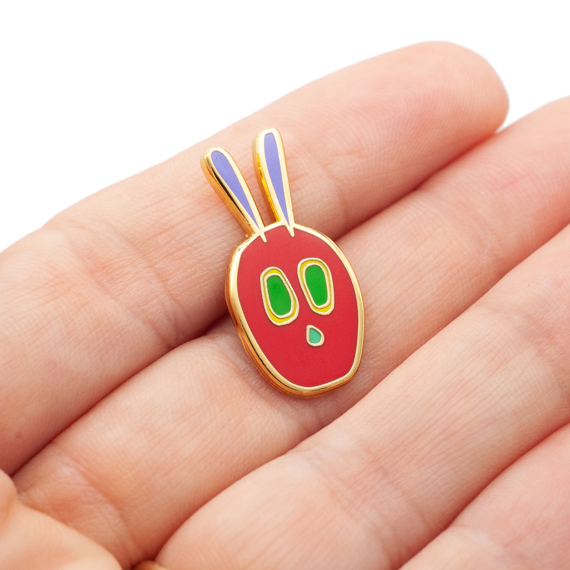 gold plated hard enamel pin with red, green, and purple fill in the shape of a caterpillar head, resting on a hand