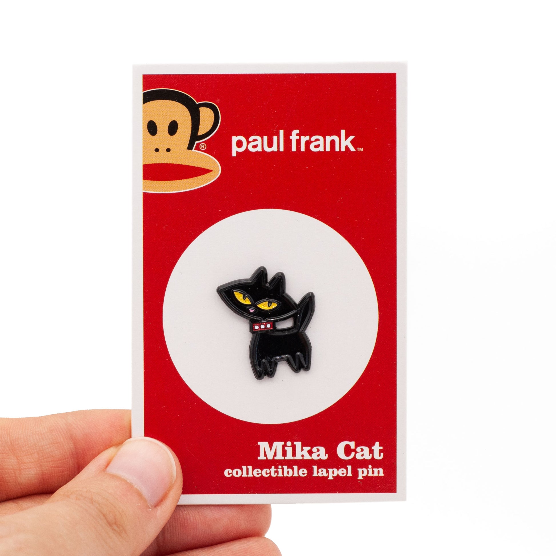 Mika Cat is a black cat enamel pin with yellow eyes and a red collar. The pin is on a backing card being held by a hand.