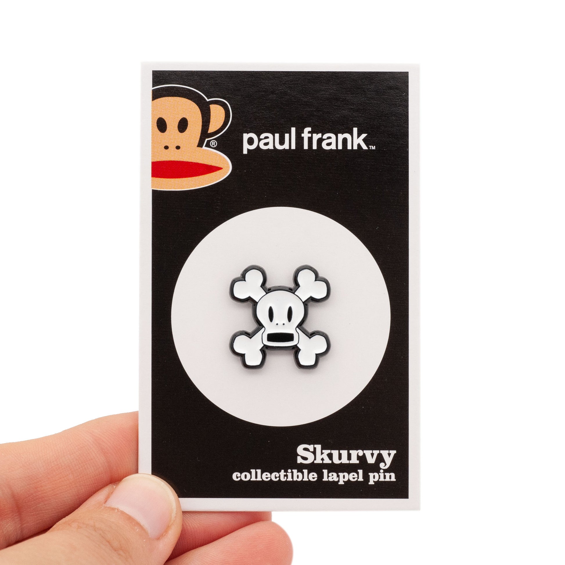 Skurvy is a soft enamel pin of white crossbones with an open mouth. shot on a backing card being held by a hand over a white background.
